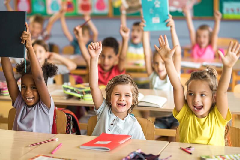 Kids smiling in class, raising their hands.